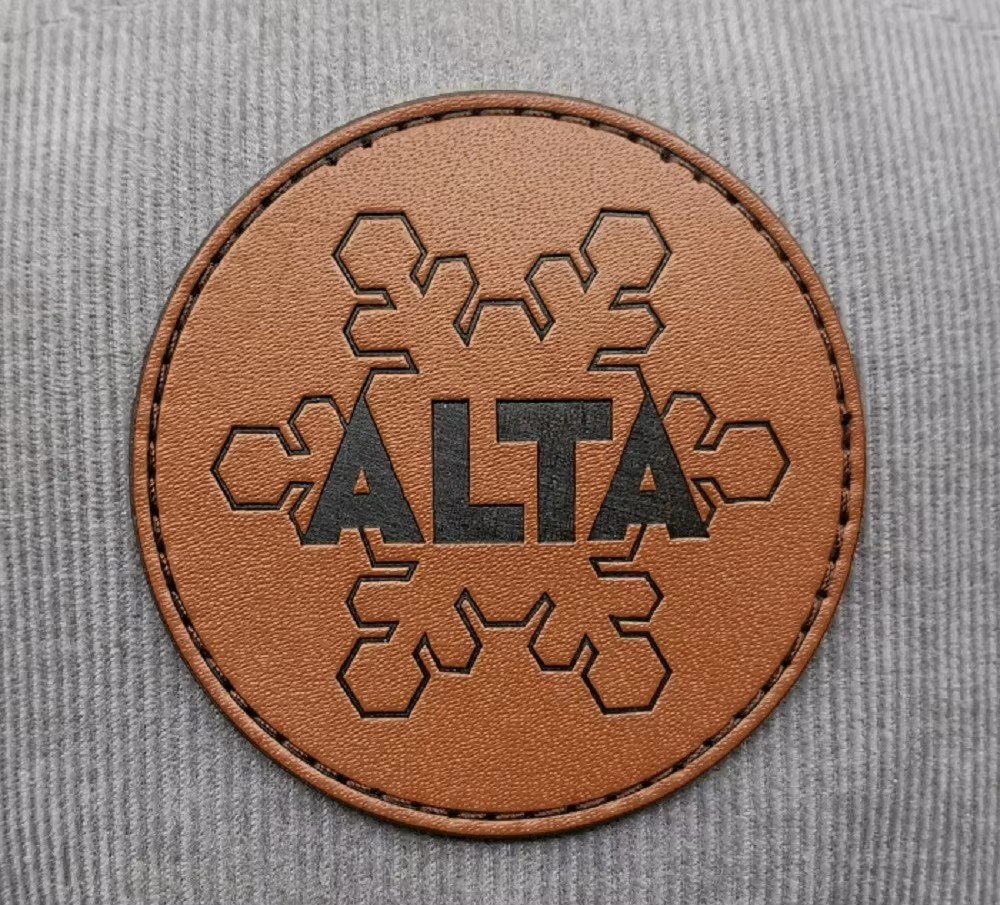 A round brown leather patch debossed with the ALTA logo, stitched onto a grey corduroy cap
