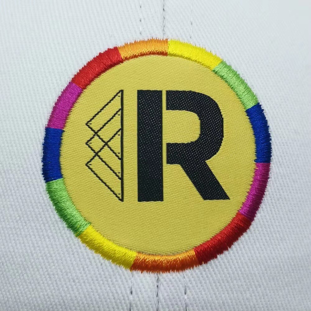 A round woven label patch with a rainbow border and black R logo on a white cap
